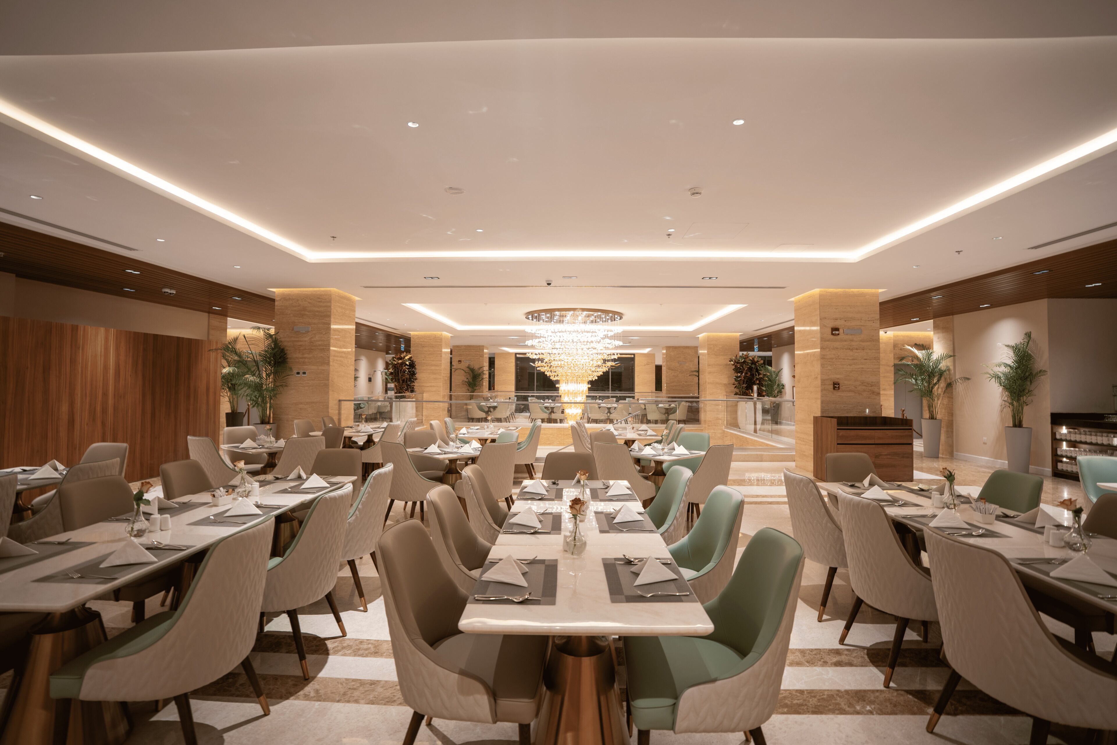 A spacious dining area with multiple tables and chairs, a central chandelier, and a view of the adjacent kitchen area.