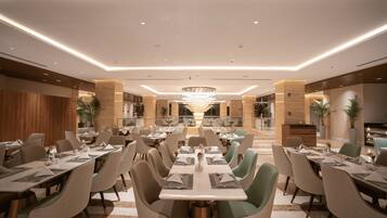 A spacious dining area with multiple tables and chairs, a central chandelier, and a view of the adjacent kitchen area.