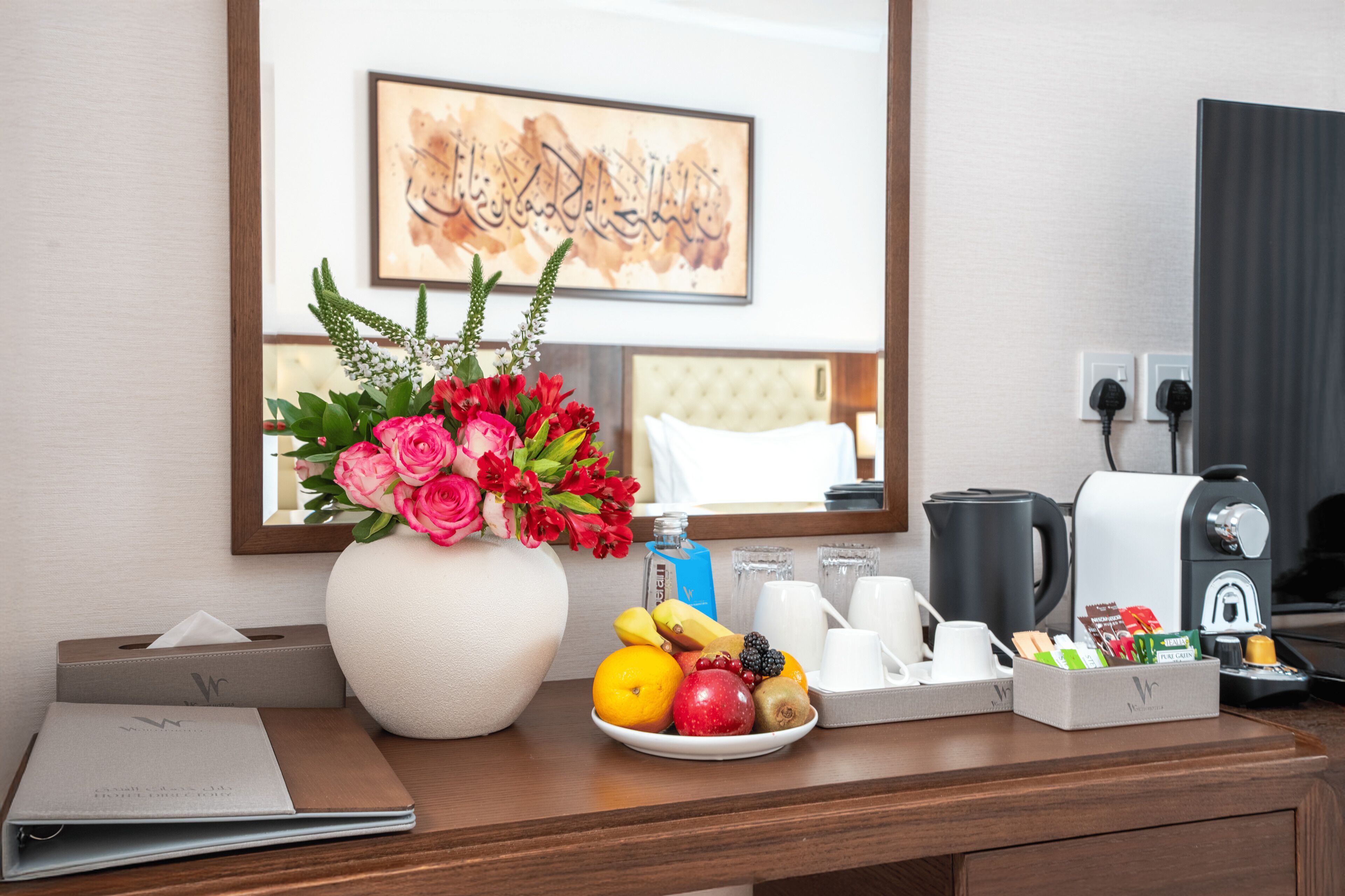A hotel room with a desk, a vase of flowers, a fruit plate, a coffee maker, and a framed artwork on the wall.