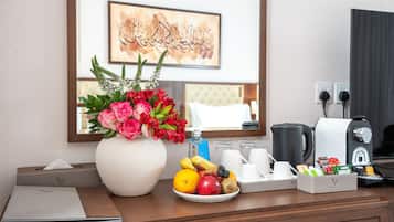 A hotel room with a desk, a vase of flowers, a fruit plate, a coffee maker, and a framed artwork on the wall.
