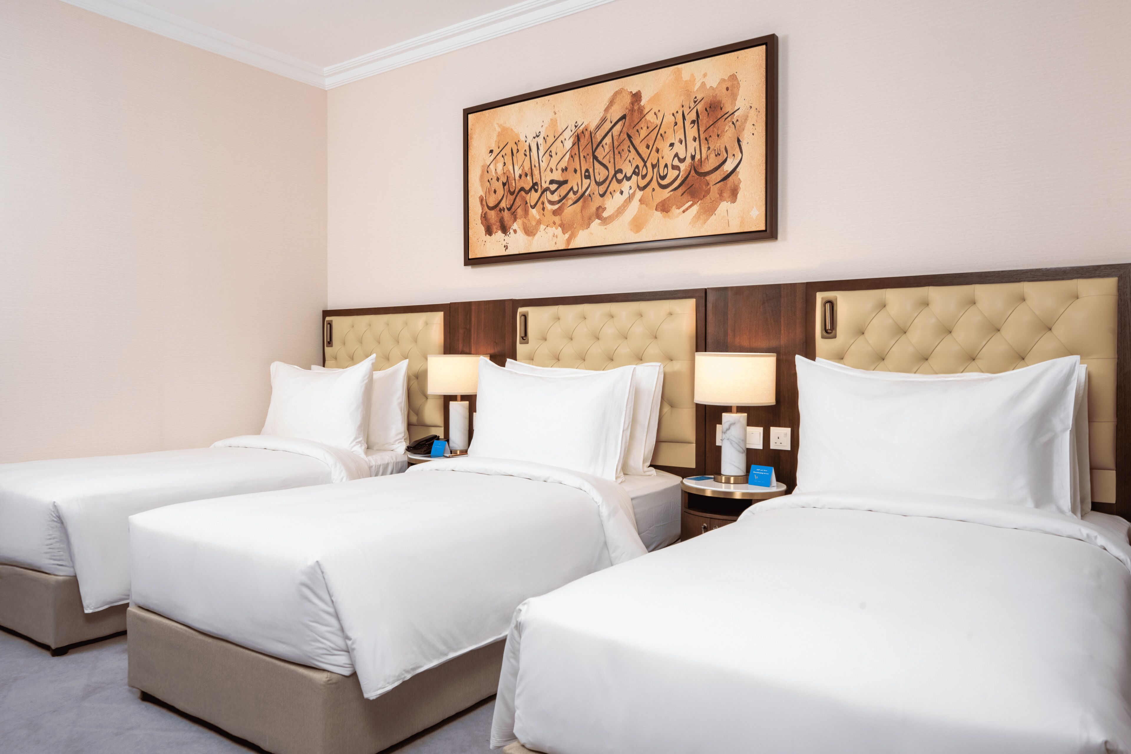 A hotel room with three beds, a wall-mounted artwork, and a nightstand with a lamp.