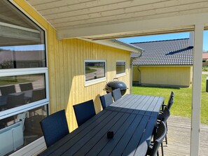 A covered outdoor dining area with a table and chairs, a grill, and a view of a residential building in the background. - Grobenbrode Escape fur 10 (Grossenbrode)