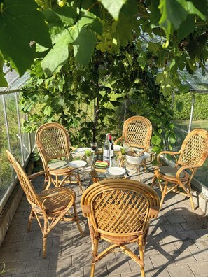 A glass-enclosed outdoor dining area with wicker chairs and a table set for a meal, surrounded by lush greenery. - Half-timbered cottage near Knuthenborg Park (Maribo)