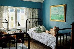 A bedroom with two beds, a window with curtains, a painting on the wall, and a lamp on the bedside table.
