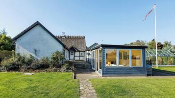 Een klassiek huis met een stenen dak, een tuinkamer met glazen deuren, en een vlaggenmast op de achtergrond.