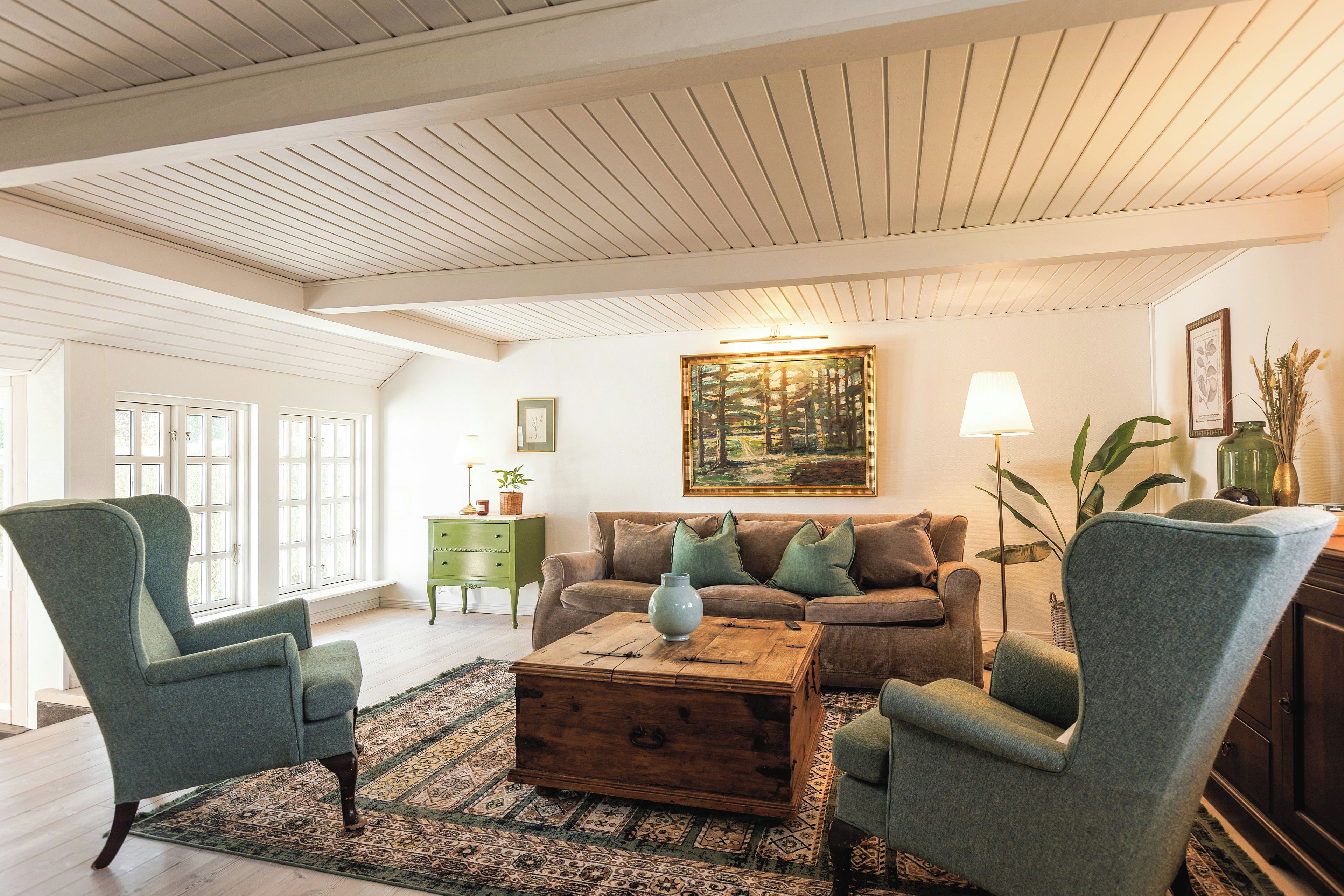 Een woonkamer met een houten plafond, een bank, twee fauteuils, een salontafel, een vloerkleed en een schilderij aan de muur.