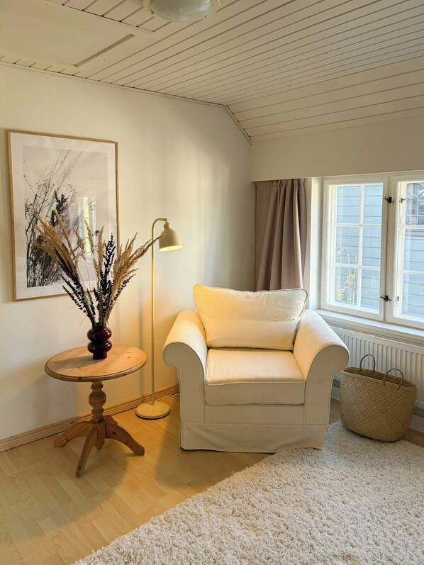 A living room with a white armchair, a round wooden table with a vase, a floor lamp, and a large window with curtains.