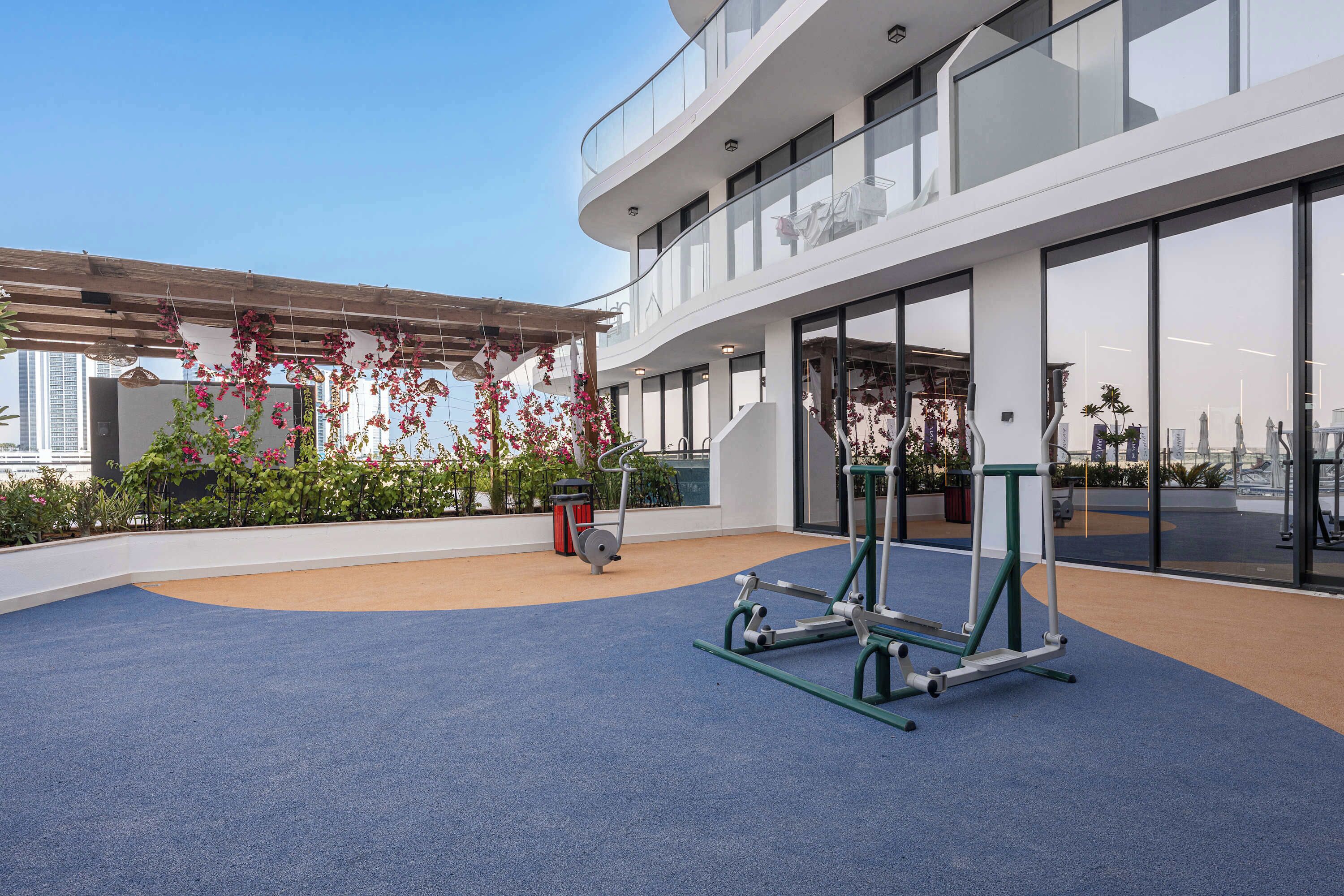 An outdoor gym area with exercise equipment, a covered seating area with plants, and a building with large windows in the background.