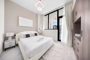 Une chambre d’hôtel moderne avec un grand lit, des tables de chevet, une petite table, une œuvre d’art accrochée au mur, une fenêtre avec des rideaux et une armoire intégrée.
