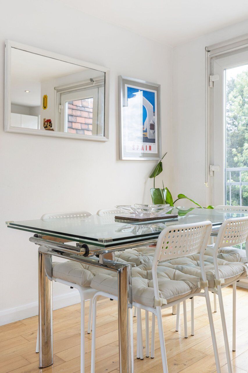 Een moderne eetruimte met een glazen tafel, witte stoelen, een grote spiegel en een ingelijst schilderij aan de wand.