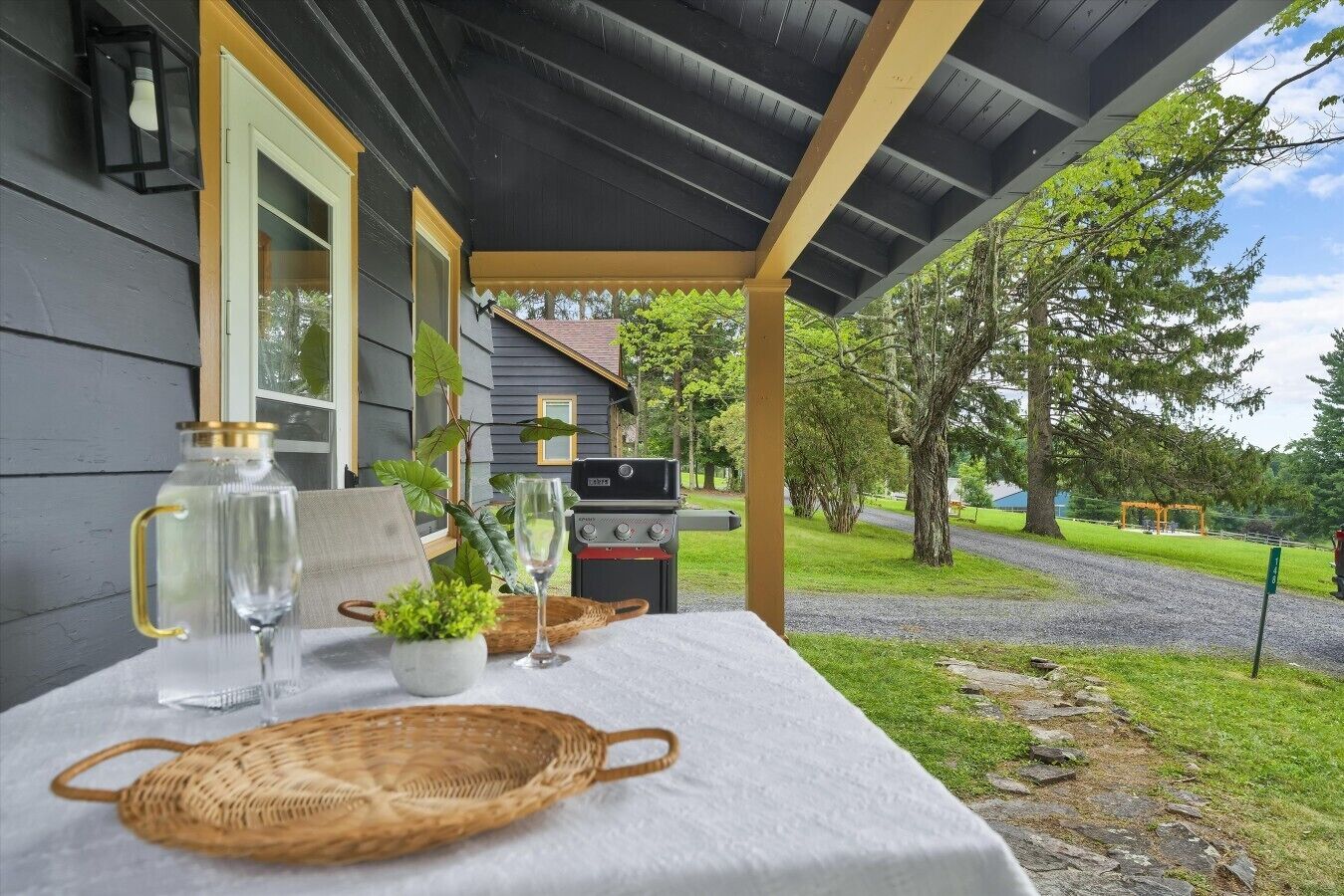 A covered outdoor seating area with a table set for two, a grill, and a view of a grassy area with trees.