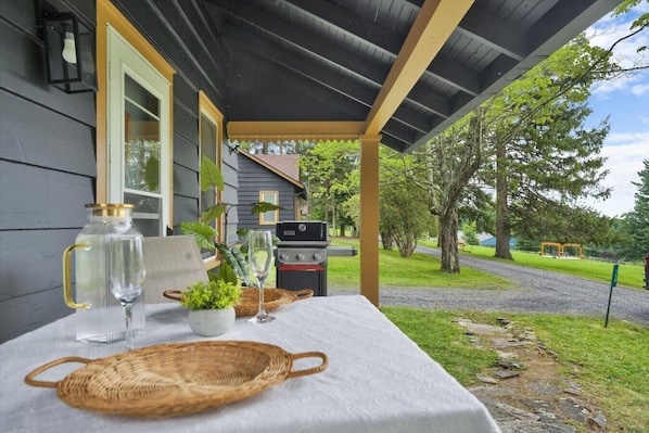 A covered outdoor seating area with a table set for two, a grill, and a view of a grassy area with trees.