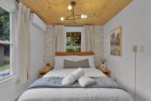 A bedroom with a wooden ceiling, a bed with white bedding, two bedside tables with lamps, a window with curtains, and a painting on the wall.