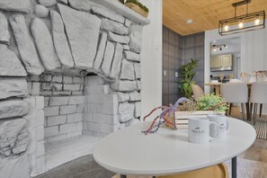 A stone fireplace, a round dining table with two white mugs, and a modern kitchen in the background.
