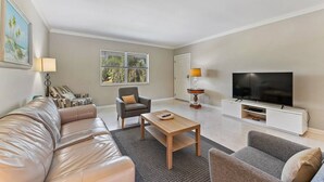 A living room with a sofa, armchair, coffee table, TV, and a window with blinds. - 2BR 2BA Updated Beach Apt with Screened Patio (West Palm Beach)