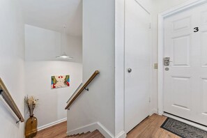 Uma escada com corrimão, uma porta com número e uma pequena obra de arte na parede.