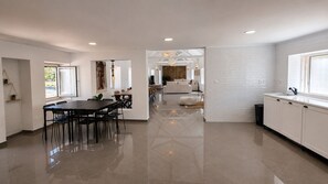 A modern kitchen with white cabinetry, a dining area with a black table and chairs, and a view into the living room. - Villa with Pool · 1 km from the Beach - 30 min from the Airport (Gaio-Rosário)