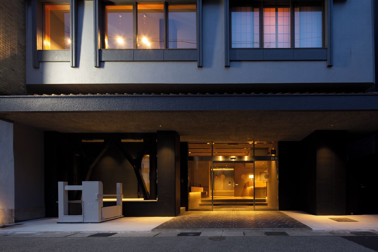 A modern building with a glass entrance, a visible staircase leading to the upper floor, and a small outdoor seating area.