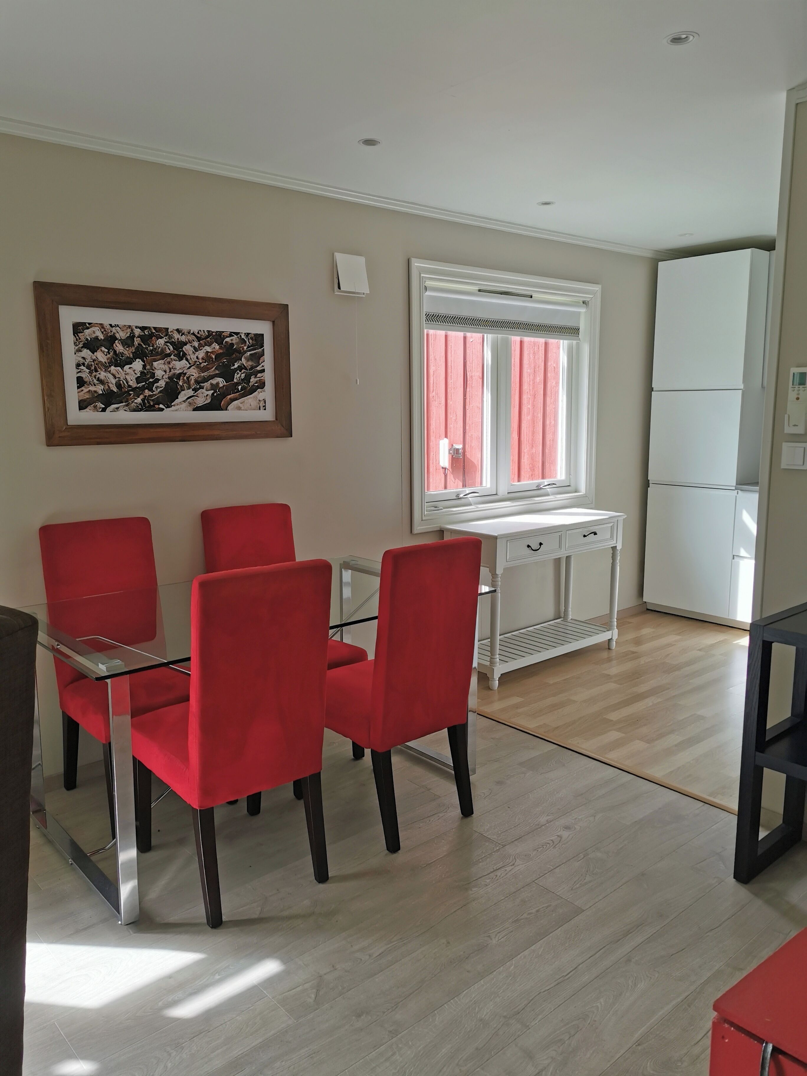Un comedor con una mesa de vidrio y sillas rojas, una ventana con cortinas, un armario blanco y un refrigerador.