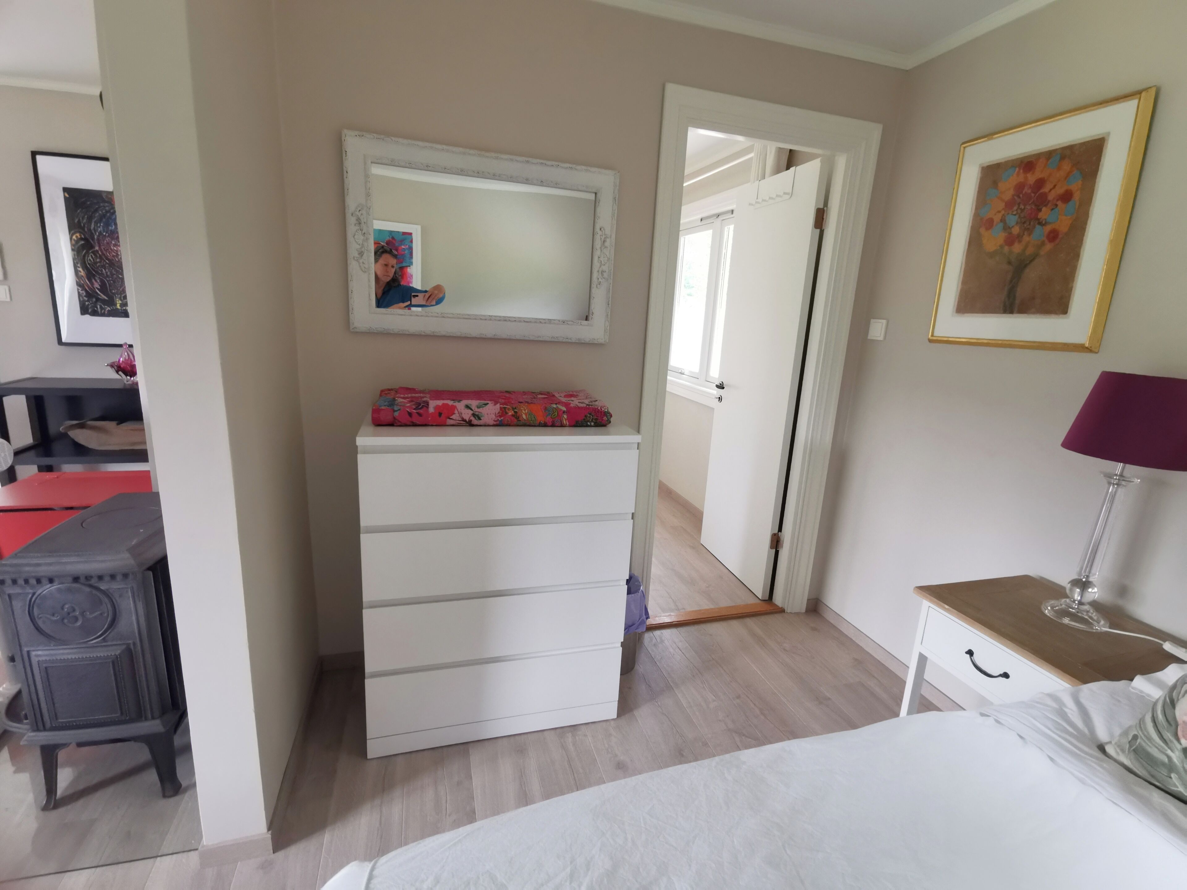 Een slaapkamer met een bed, een dressoir, een spiegel, een lamp en een deur naar een andere kamer.