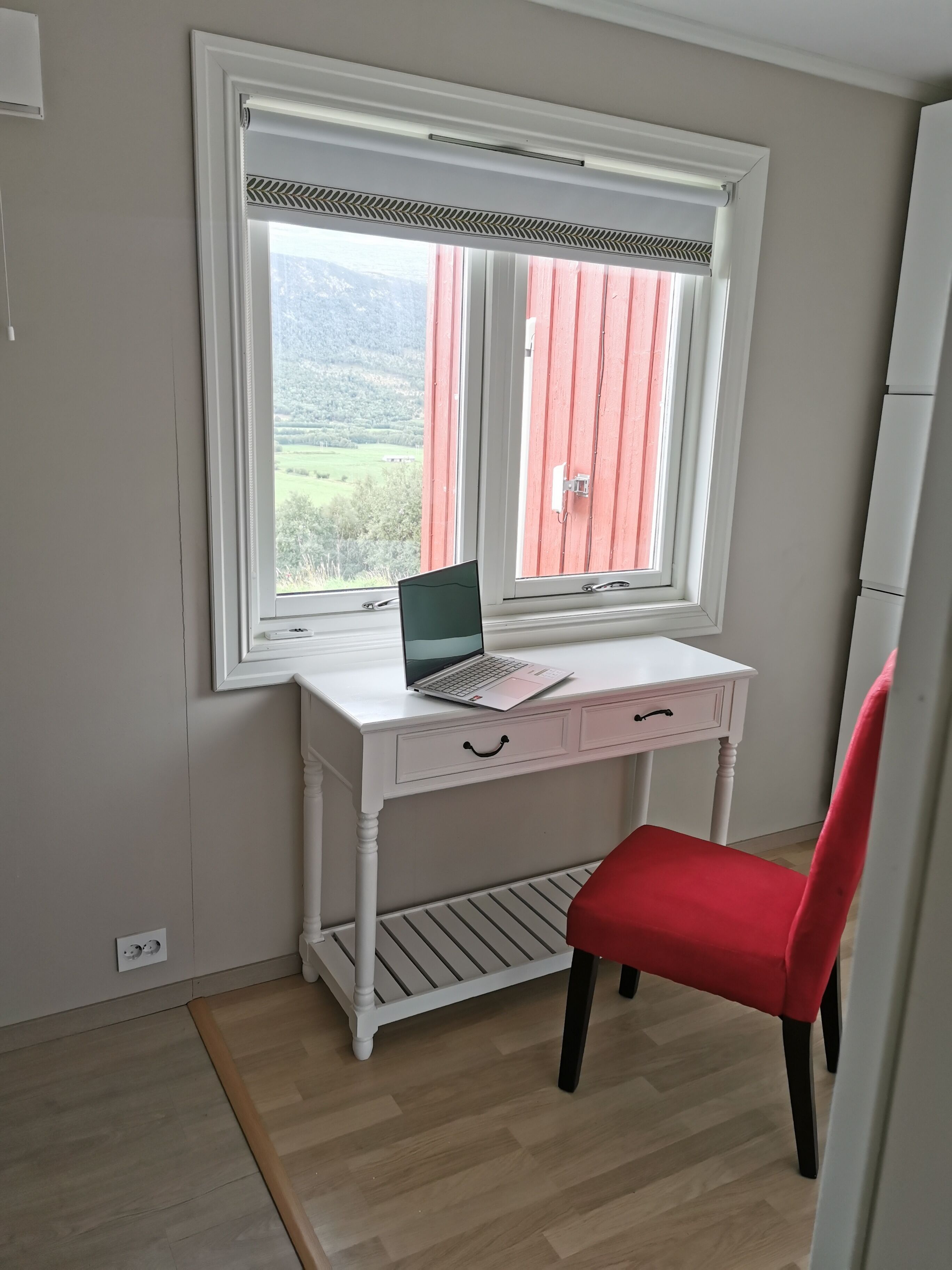 A room with a white desk, a red chair, and a window with a view of greenery and hills.