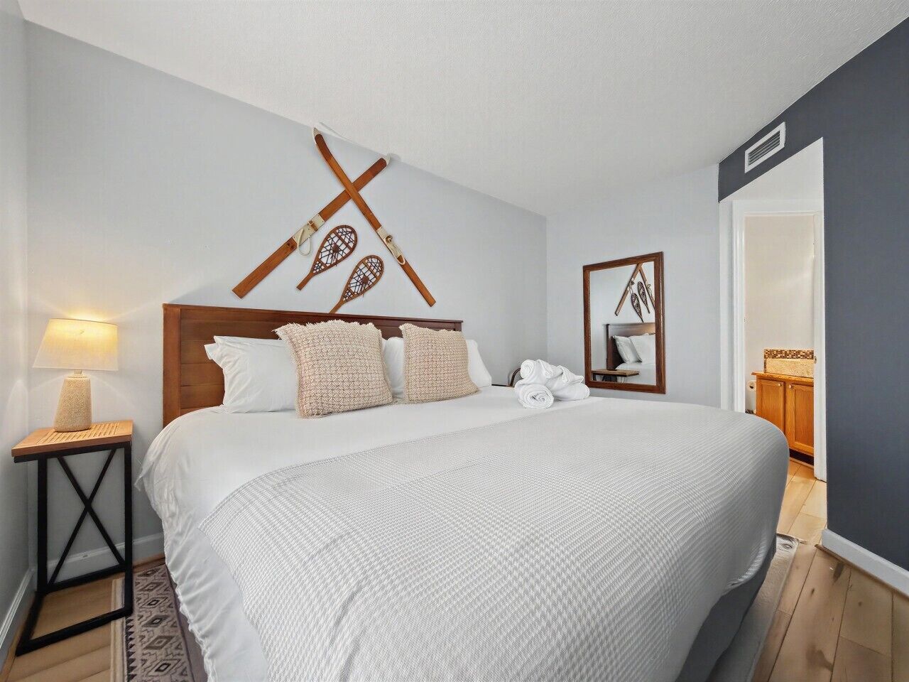 A neatly made bed with a white bedspread and pillows, a wooden headboard, a bedside table with a lamp, and a wall-mounted mirror.