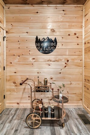 Ein Holzblockhaus-Interieur mit einem Schild, auf dem steht 'DIE BERGE rufen und ich muss hin', sowie ein Wagen mit Gläsern und einer Pflanze.