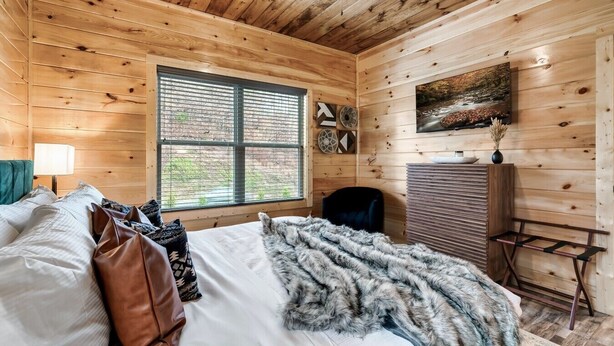 Camera da letto in una baita di legno con un letto, cuscini, una coperta, una sedia, una scrivania e una finestra con persiane.