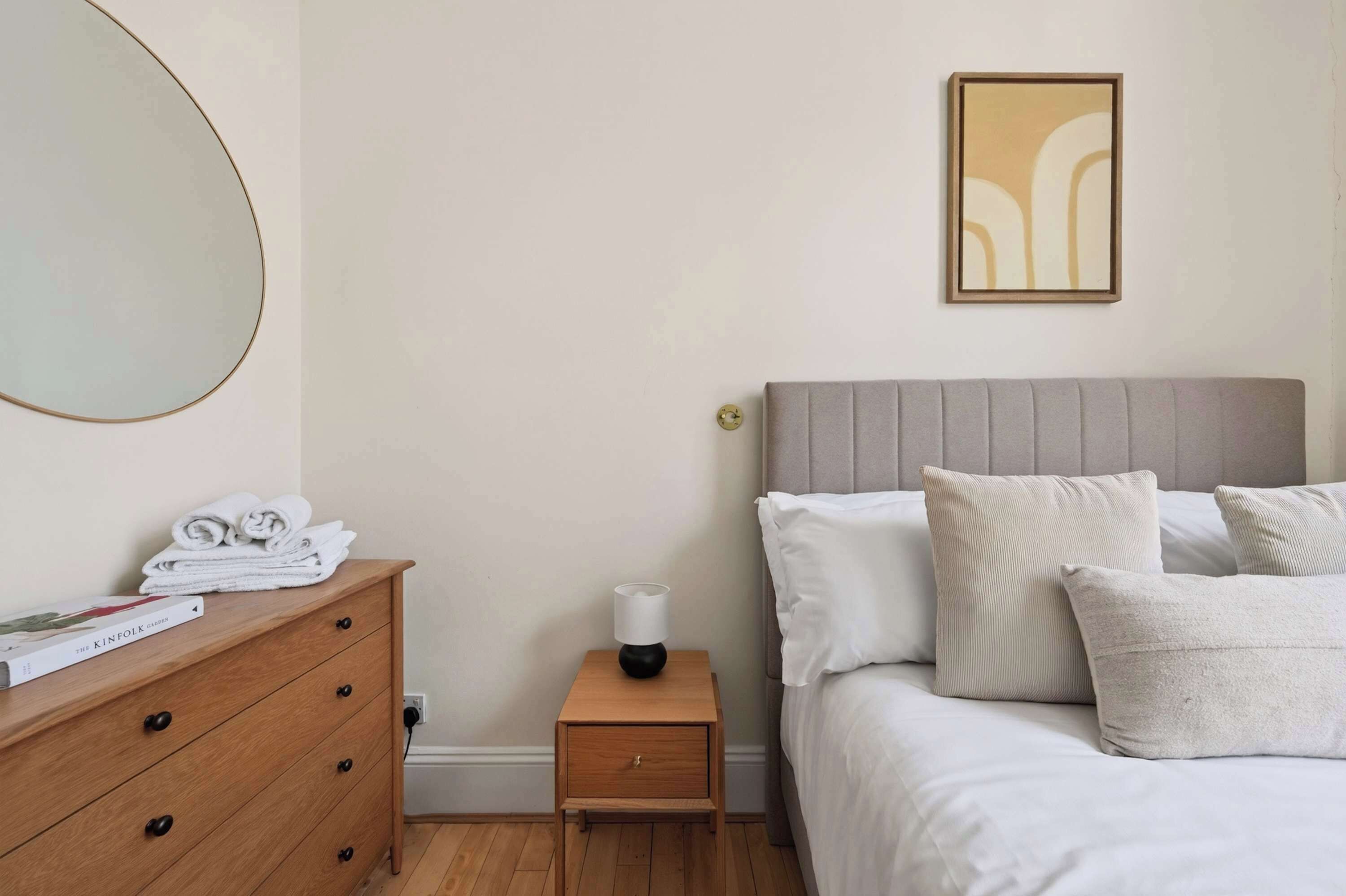 Un dormitorio con cama, mesita de noche con lámpara, cómoda con toallas y un cuadro en la pared.