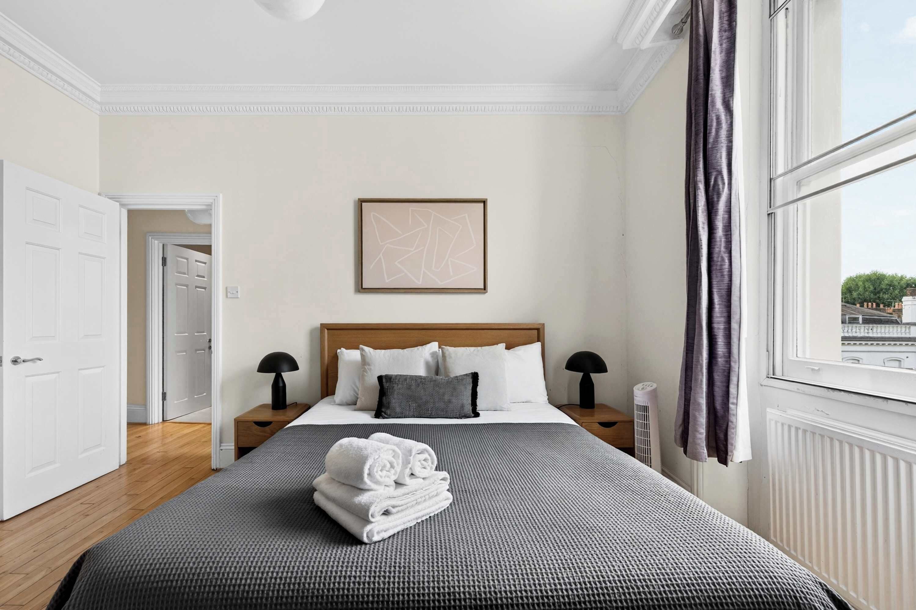 A hotel room with a large bed, a bedside table, a nightstand, a framed picture on the wall, and a window with curtains.