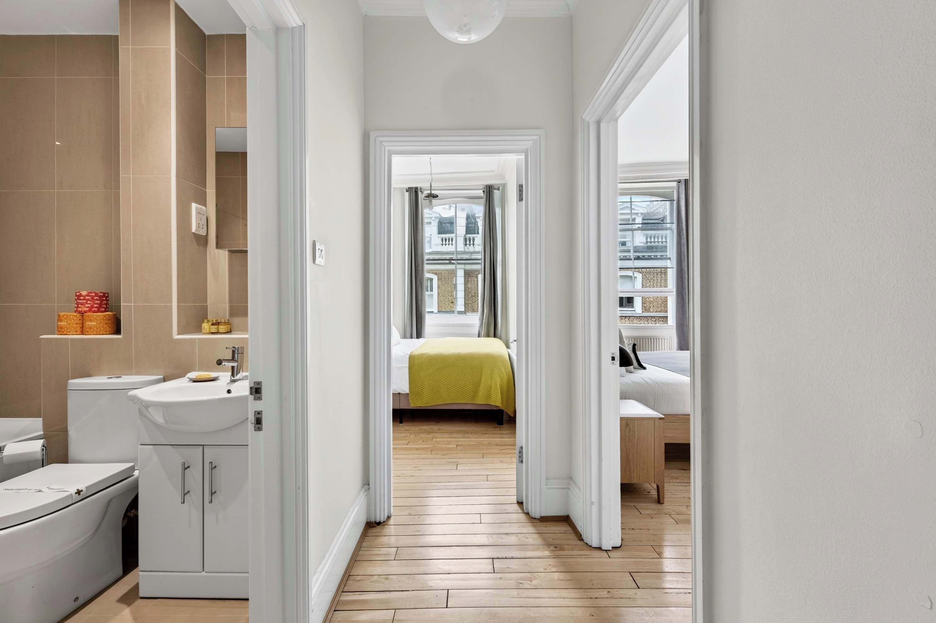 Een smalle gang die leidt naar een slaapkamer met een bed, een badkamer met een wastafel, en uitzicht op het omliggende landschap.