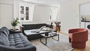 Ein modernes Wohnzimmer mit einer Couch, einem Couchtisch und einem roten Sessel. Es gibt einen Essbereich mit Tisch und Stühlen sowie eine Küche mit einer Waschmaschine.