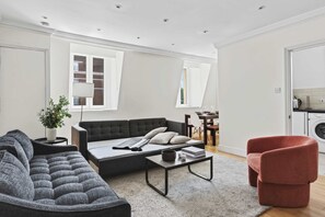 Ein modernes Wohnzimmer mit einer Couch, einem Couchtisch und einem roten Sessel. Es gibt einen Essbereich mit Tisch und Stühlen sowie eine Küche mit einer Waschmaschine.