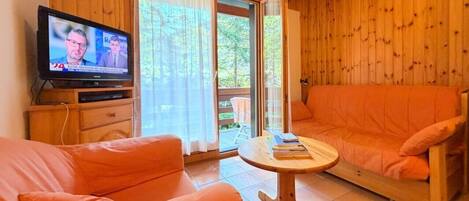 A room with a wooden interior, a television on a stand, an orange sofa, and a small round table with books.