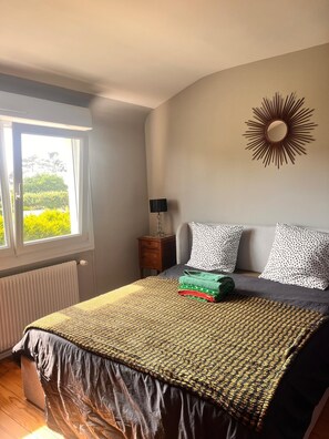 Ein Schlafzimmer mit einem Bett, einem Nachttisch, einer Lampe, einem Fenster mit Blick auf Grünflächen und einem Sonnenspiegel an der Wand.
