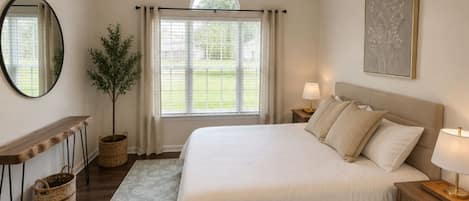 A bedroom with a bed, bedside tables, a ceiling fan, a window with curtains, a mirror, a potted plant, and a rug.