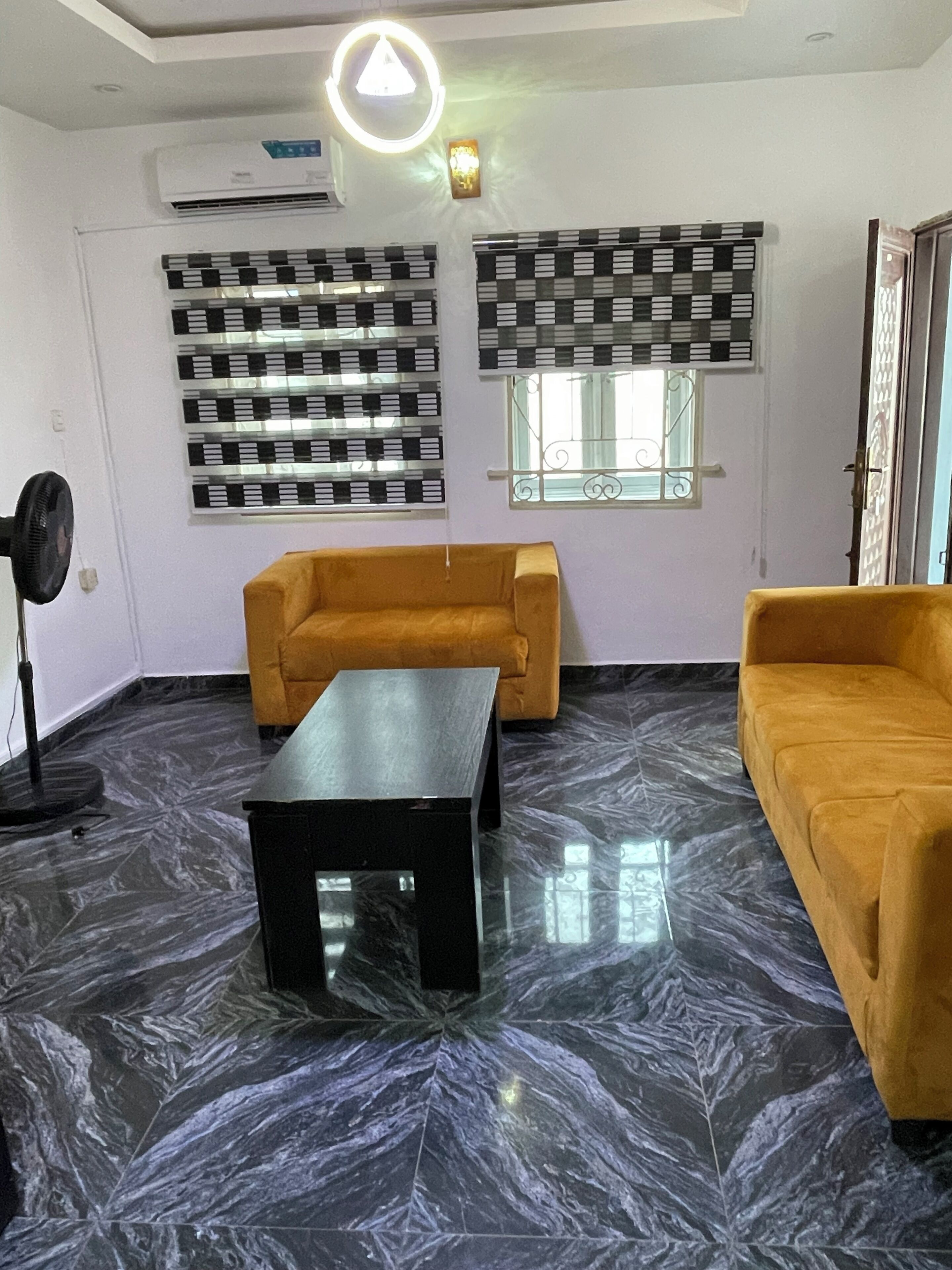 Une pièce comprenant un canapé, une table basse, un ventilateur sur pied et une fenêtre agrémentée de stores décoratifs.
