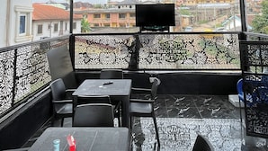 Un área de estar al aire libre con techo, mesas y sillas, barandilla con diseño y vista de edificios al fondo.