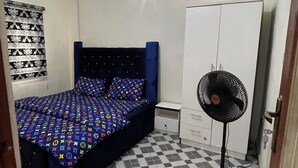 Un dormitorio con piso a cuadros, una cama con colcha estampada, un ventilador de pie y un armario.