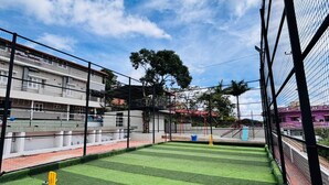 Cancha de cricket interior con césped artificial, rodeada por una estructura de malla y un edificio de varios pisos al fondo.