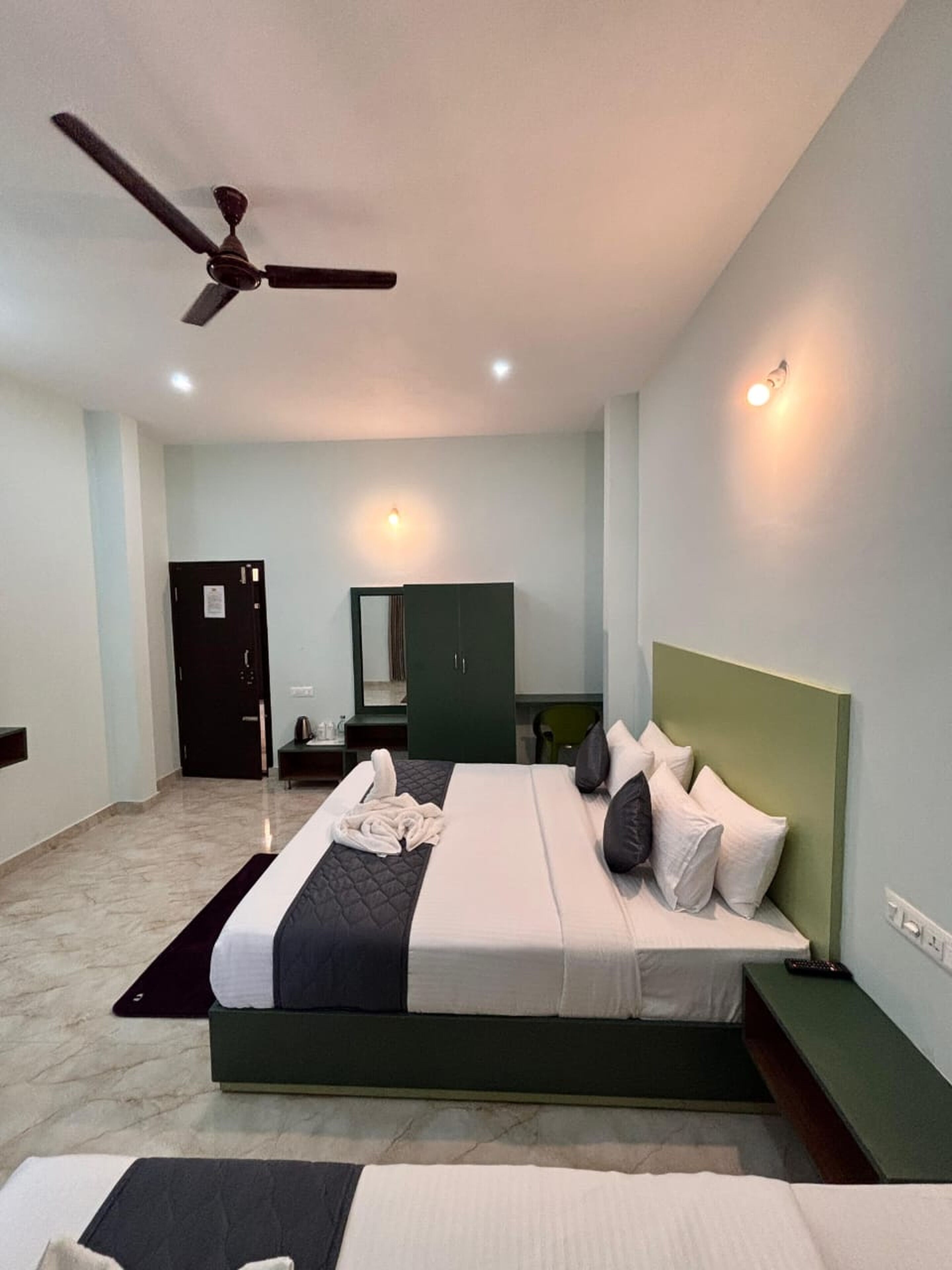 A hotel room with a bed, a desk, a chair, a wardrobe, a ceiling fan, and a door.