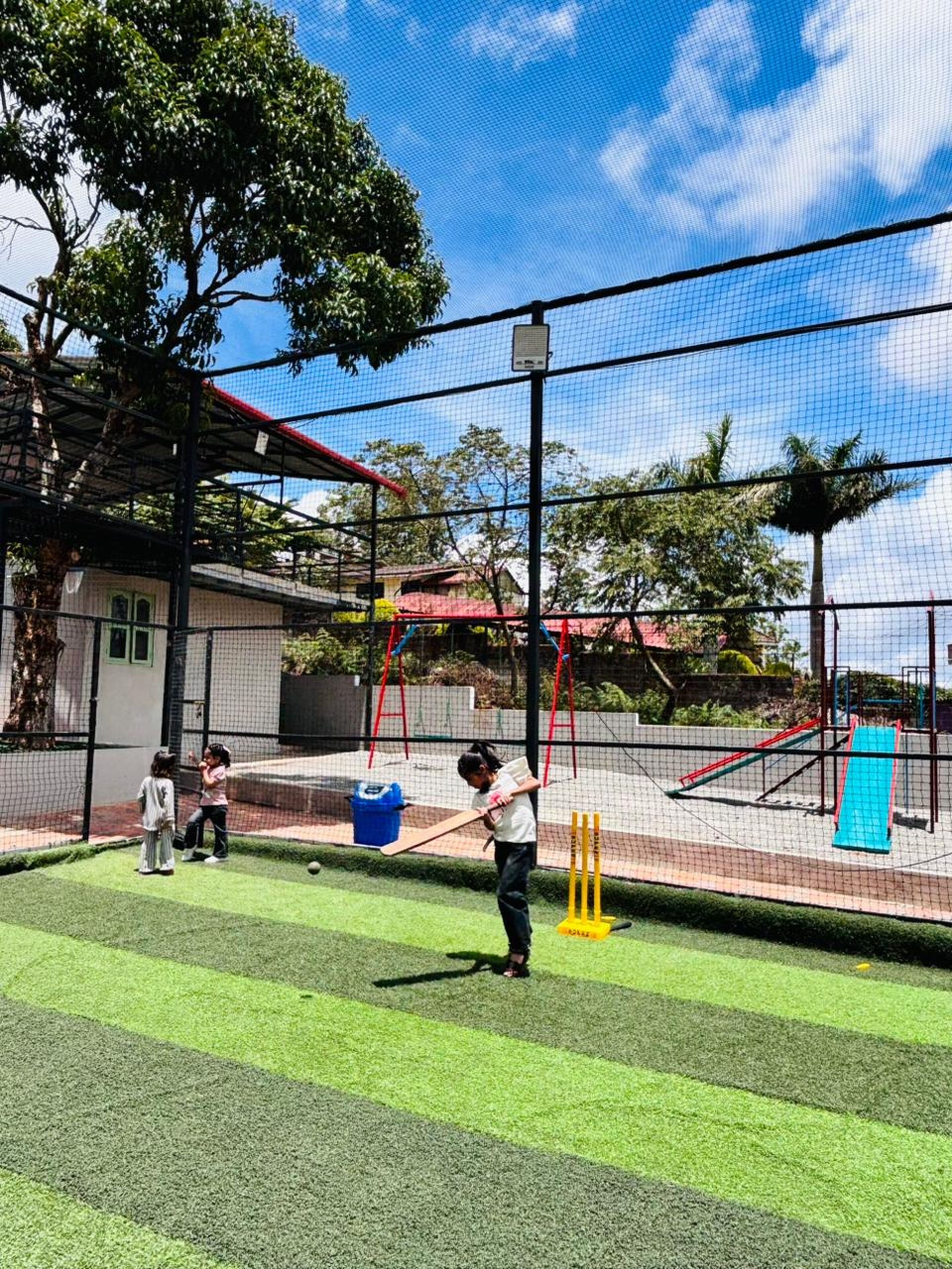 Zona de entrenamiento de críquet al aire libre con una valla de malla, un jaulón para bateadores y un pequeño pabellón.