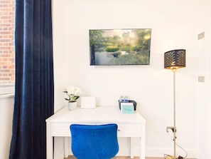 Una scrivania bianca con una sedia blu, un quadro incorniciato di un paesaggio, un vaso con fiori bianchi e un cartello di benvenuto.
