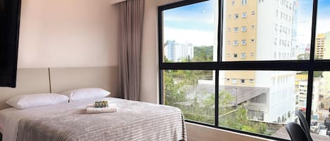 A hotel room with a large window, a bed with white linens, and a view of city buildings.