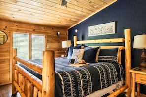 Een slaapkamer in chaletstijl met een bed, kussens, een nachtkastje en een wandklok.