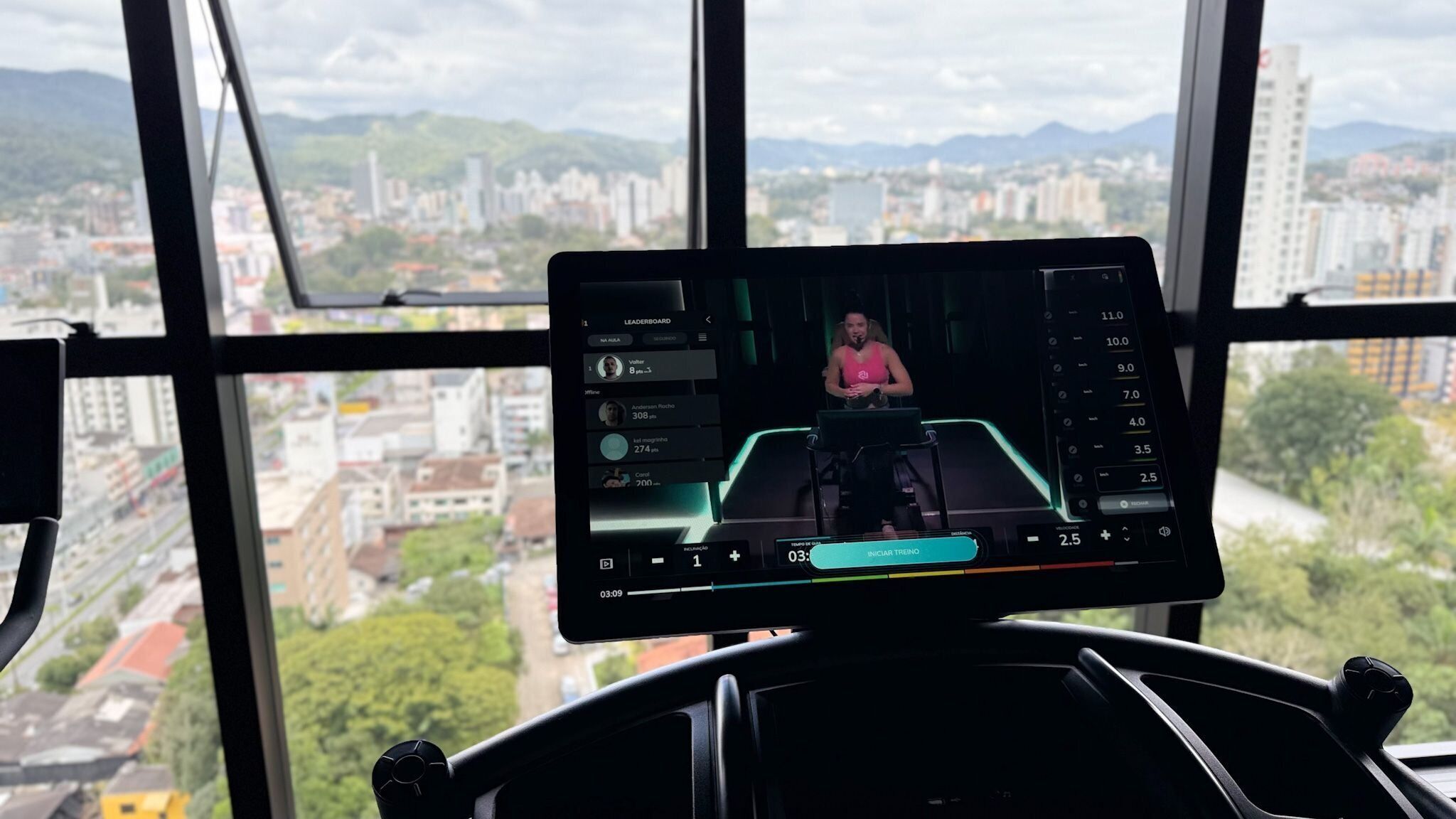 Una cinta de correr con pantalla digital, vistas panorámicas de la ciudad a través de amplias ventanas y una persona haciendo ejercicio.