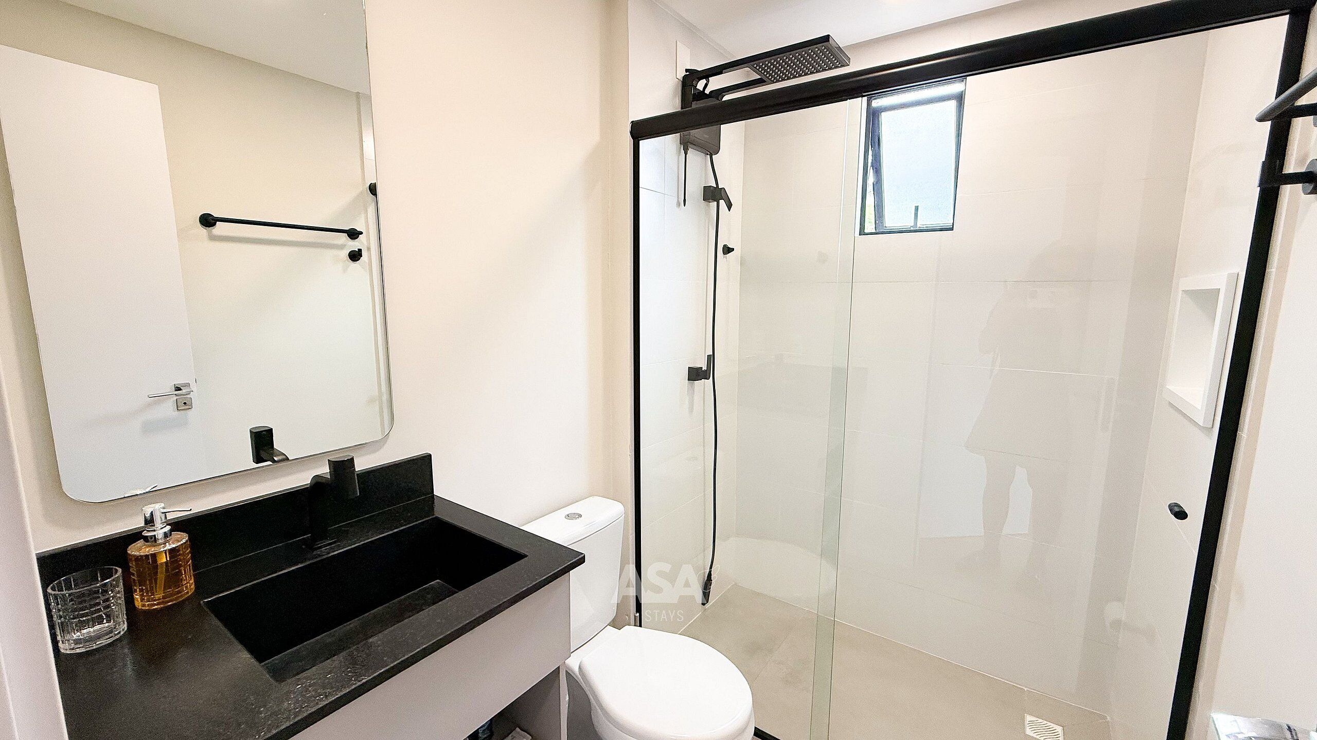 A bathroom with a glass shower enclosure, a white toilet, a black sink with a soap dispenser, and a mirror above the sink.