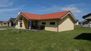 Una casa amarilla con techo rojo, un patio con sillas y un cielo azul despejado.