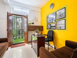 A room with a yellow wall, a wooden door, a desk with a plant, a black chair, and a couch. - Deluxe Cochin Apartments Annex (Aluva)