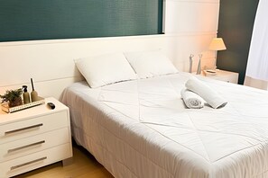 Una cama bien hecha con un edredón blanco y almohadas, una mesita de noche con una lámpara, una planta pequeña y una toalla enrollada.